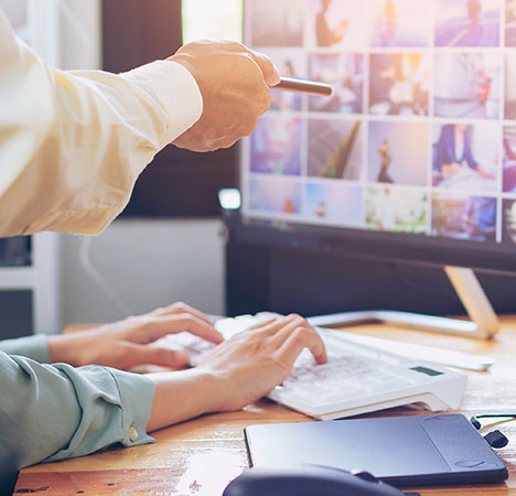 Zwei Hände auf einer Tastatur, eine dritte zeigt mit einem Stift auf einen Monitor